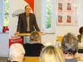 Klaus Grothe-Bortlik steht an einem Rednerpult und spricht zu einem Publikum. Im Hintergrund ist das Logo des Selbsthilfezentrums zu sehen